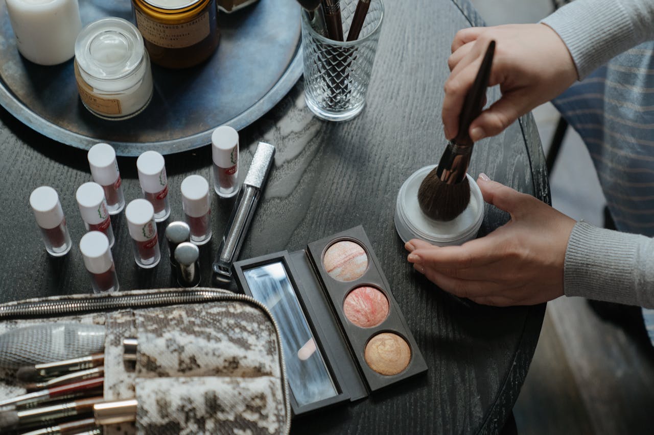 A flatlay of makeup and beauty products including eyeshadows, lipsticks, and brushes on a table.